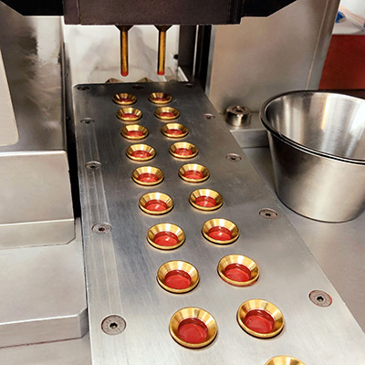 Maschine füllt rote Lippenstiftmasse in goldfarbene Negativformen auf einer Metallplatte ab