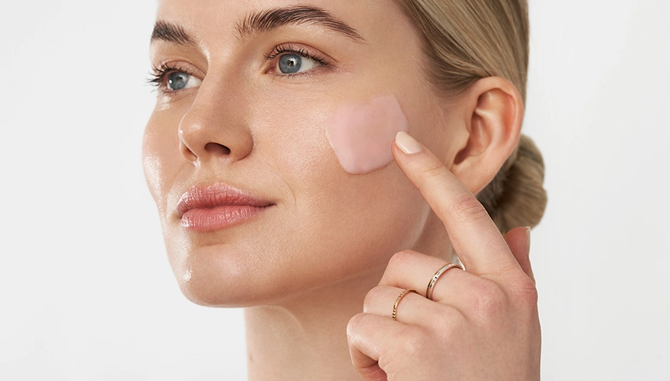 Hautalterung vorbeugen Frau trägt mit Finger rosa Creme auf die rechte Wange auf, neutraler Hintergrund, Nahaufnahme