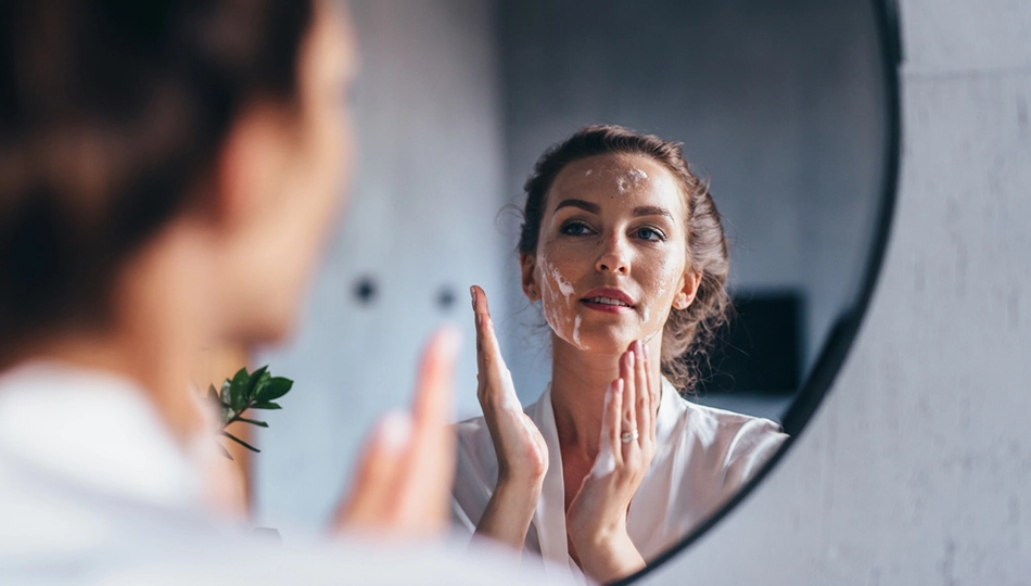 Naturkosmetik Unreine Haut Frau vor dem Badezimmerspiegel, trägt Alltags-Make-up und reinigt ihr Gesicht mit mildem Reinigungsschaum.