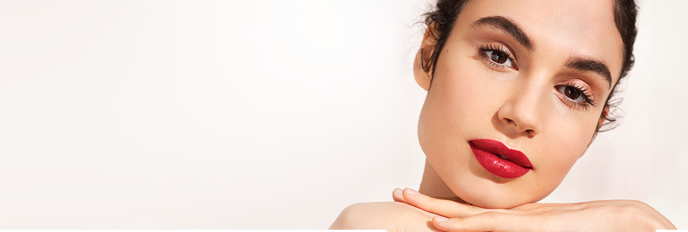 Nahaufnahme einer Frau mit roten Lippen, ruhiger Mimik und Händen unter dem Kinn vor hellem Hintergrund