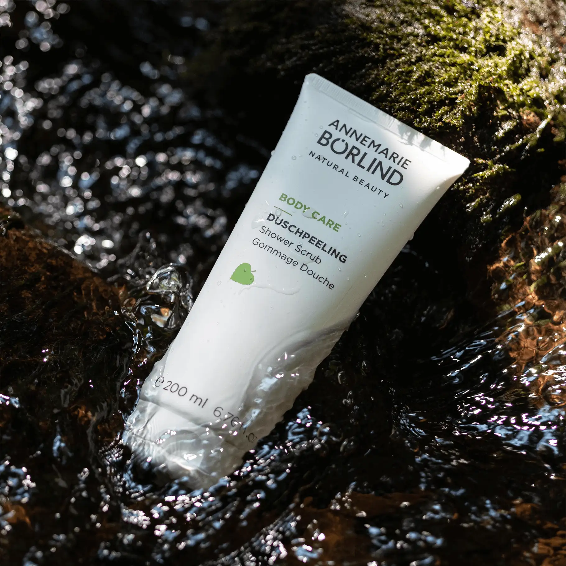 ANNEMARIE BÖRLIND Body Care Duschpeeling in weißer Tube liegt im fließenden Wasser auf Moos
