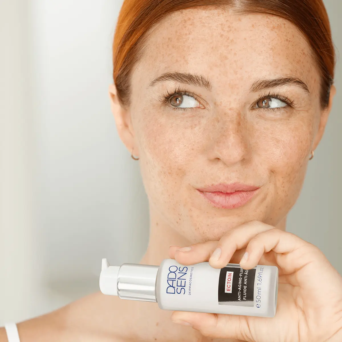 Frau hält DADO SENS ECTOIN Anti-Aging-Fluid 50 ml, Blick nach rechts, helle Haut mit Sommersprossen