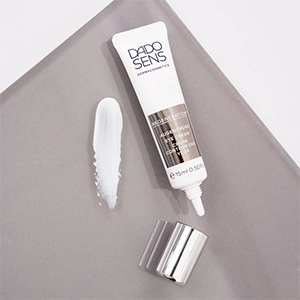 Augencreme für empfindliche Augen: DADO SENS REGENERATIONE Augencreme mit abgenommener Verschlusskappe und einem Klecks cremiger Textur, inszeniert auf grauem Untergrund.