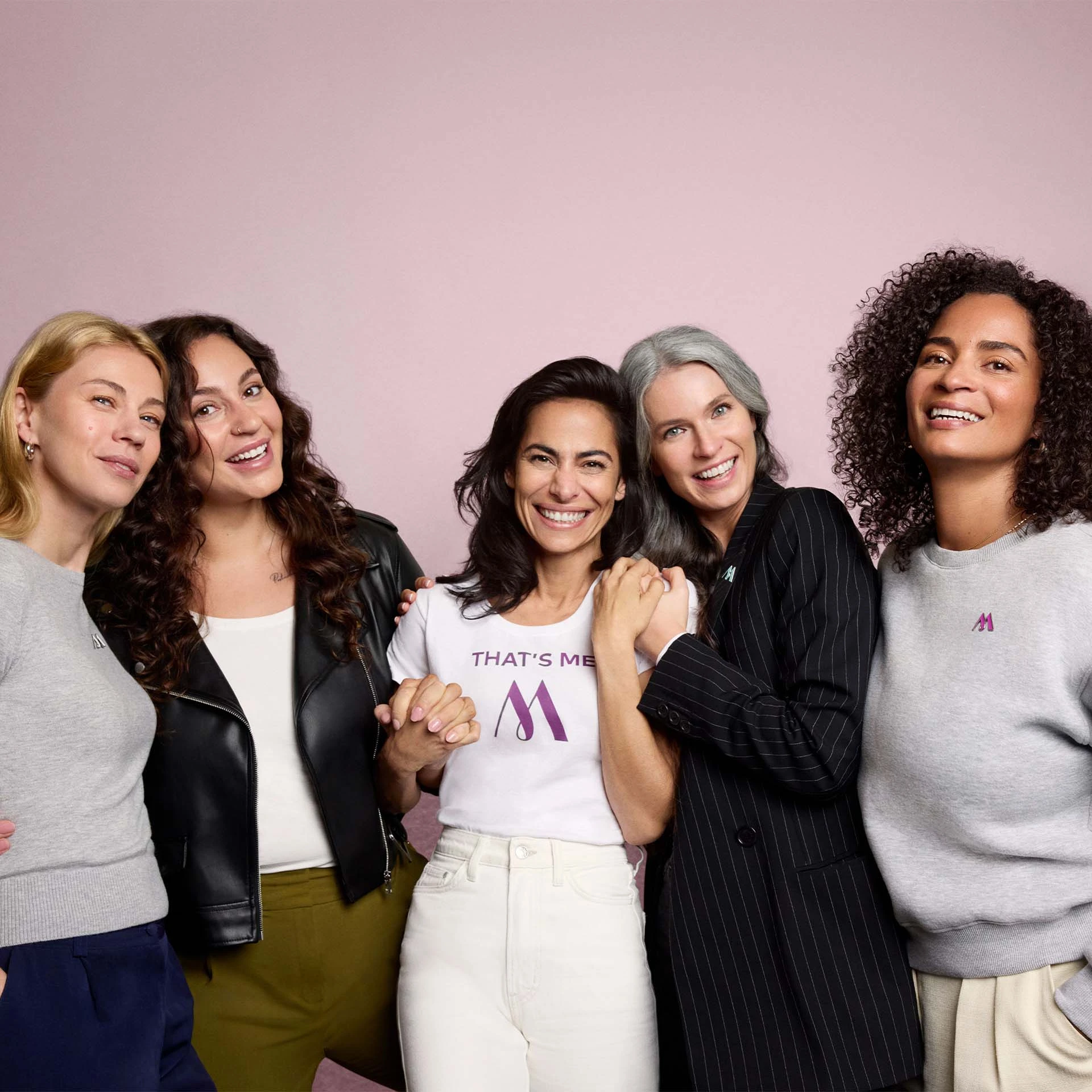 Fünf Frauen verschiedener Altersgruppen lachen und halten sich vor rosa Hintergrund an den Händen