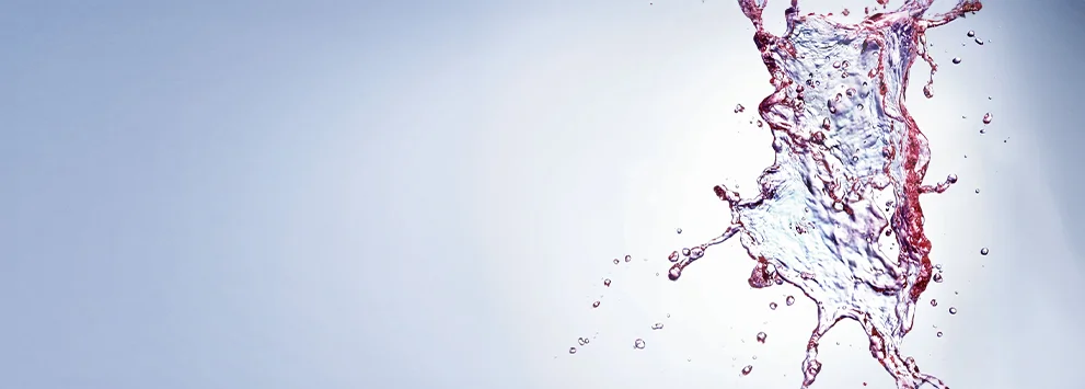 Spritzendes Wasser mit roten Farbtropfen vor hellem, unscharfem Hintergrund, dynamische Bewegung