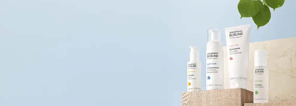 ANNEMARIE BÖRLIND Körperpflege mit Körperöl, Duschschaum, Bodylotion und Deo Spray auf Holzpodest vor blauem Hintergrund