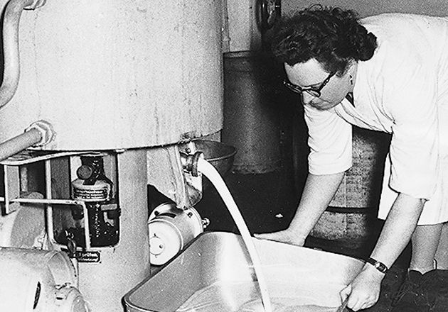 Frau in Arbeitskleidung füllt Behälter mit Flüssigkeit aus industriellem Metallbehälter in Schwarz-Weiß-Foto