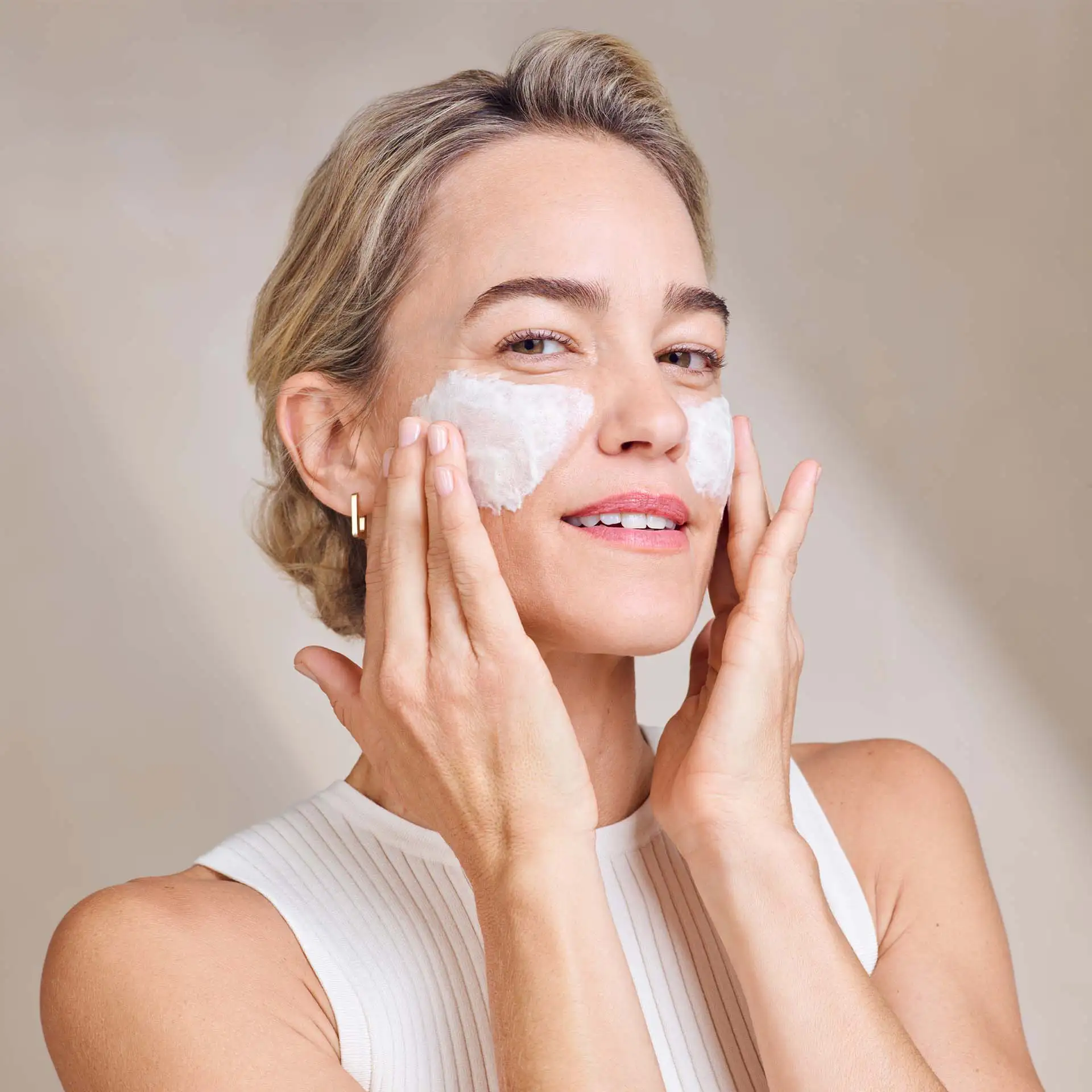 Frau trägt cremigen Cleanser auf beide Wangen auf, helle Kleidung, neutraler Hintergrund