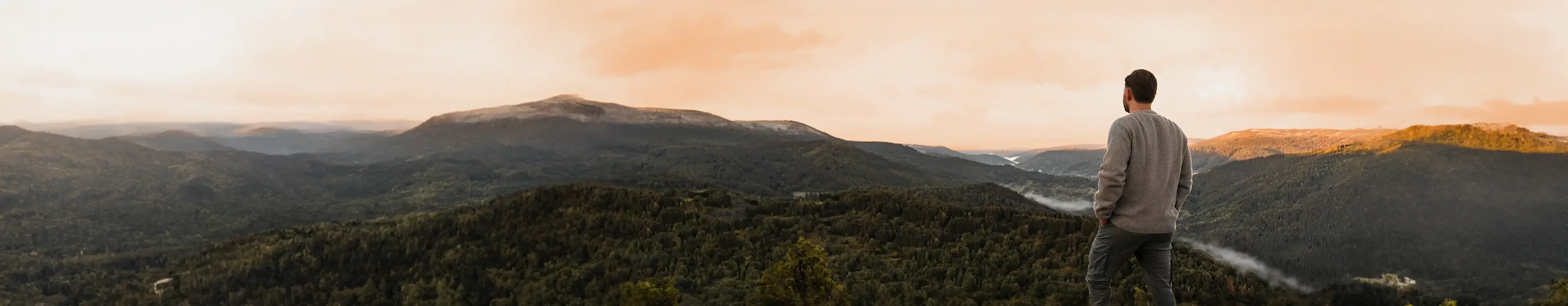 Mann in grauem Pullover blickt auf bewaldete Berge bei Sonnenuntergang mit orangefarbenem Himmel