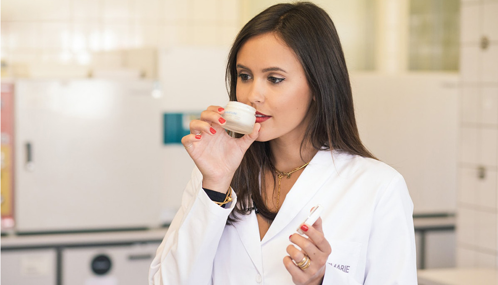 Frau in weißem Kittel riecht an einem kleinen Tiegel mit Hautpflegeprodukt in einem Laborraum