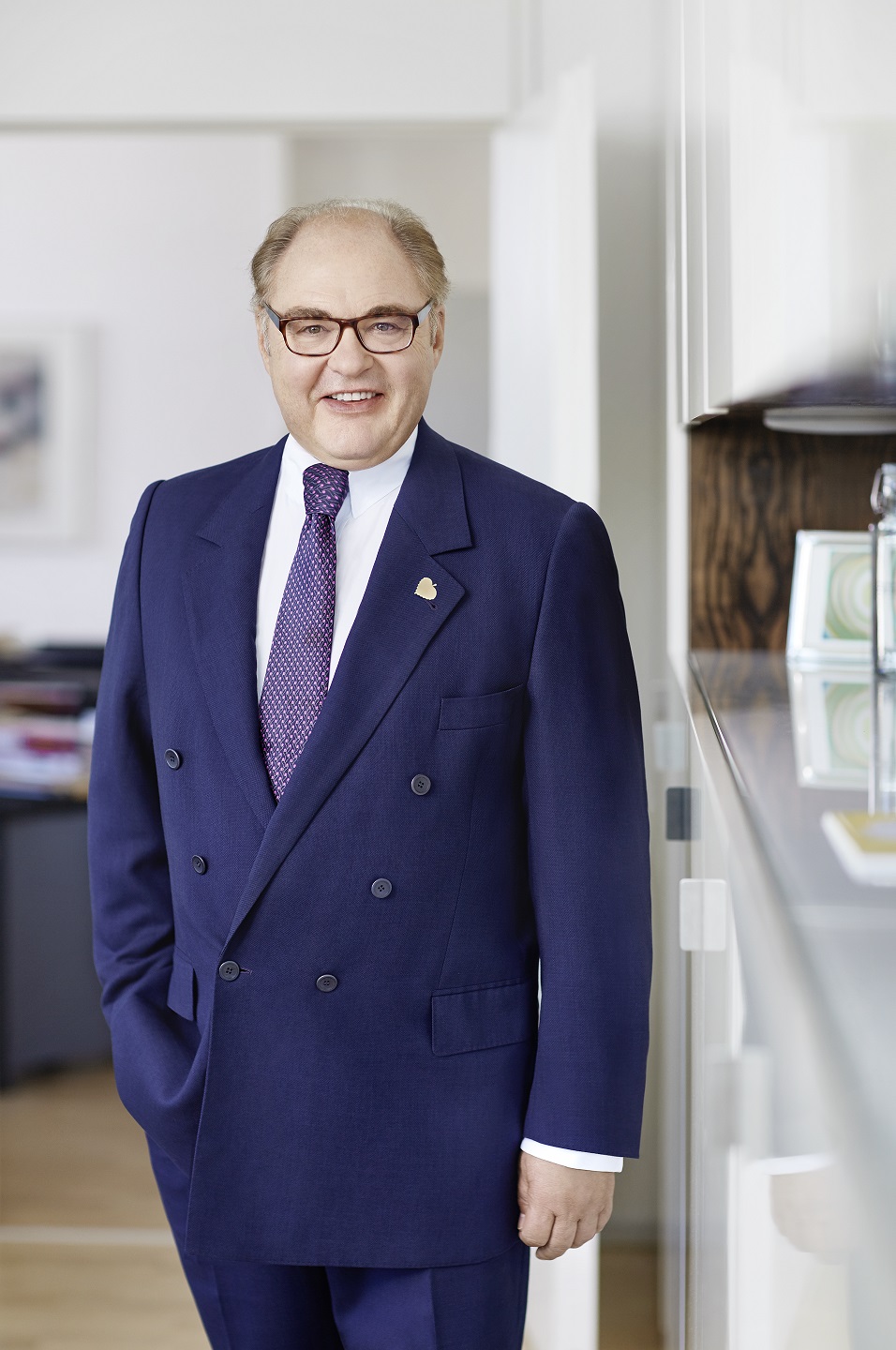 Michael Lindner in blauem Anzug und Krawatte steht lächelnd in einem hellen Büro mit Händen in den Taschen