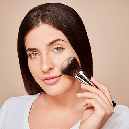 Frau trägt mit Pinsel Puder auf die rechte Wange auf, neutraler Hintergrund, natürliche Make-up-Situation