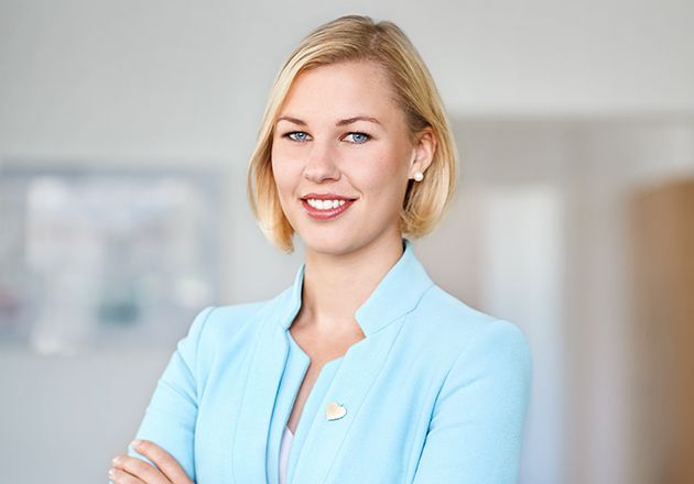 Frau in hellblauem Blazer steht mit verschränkten Armen und lächelt in einem hellen Büro