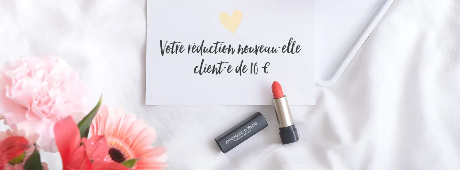 ANNEMARIE BÖRLIND Lippenstift in Korallrot neben Karte mit französischem Text auf weißem Stoff, Blumen links
