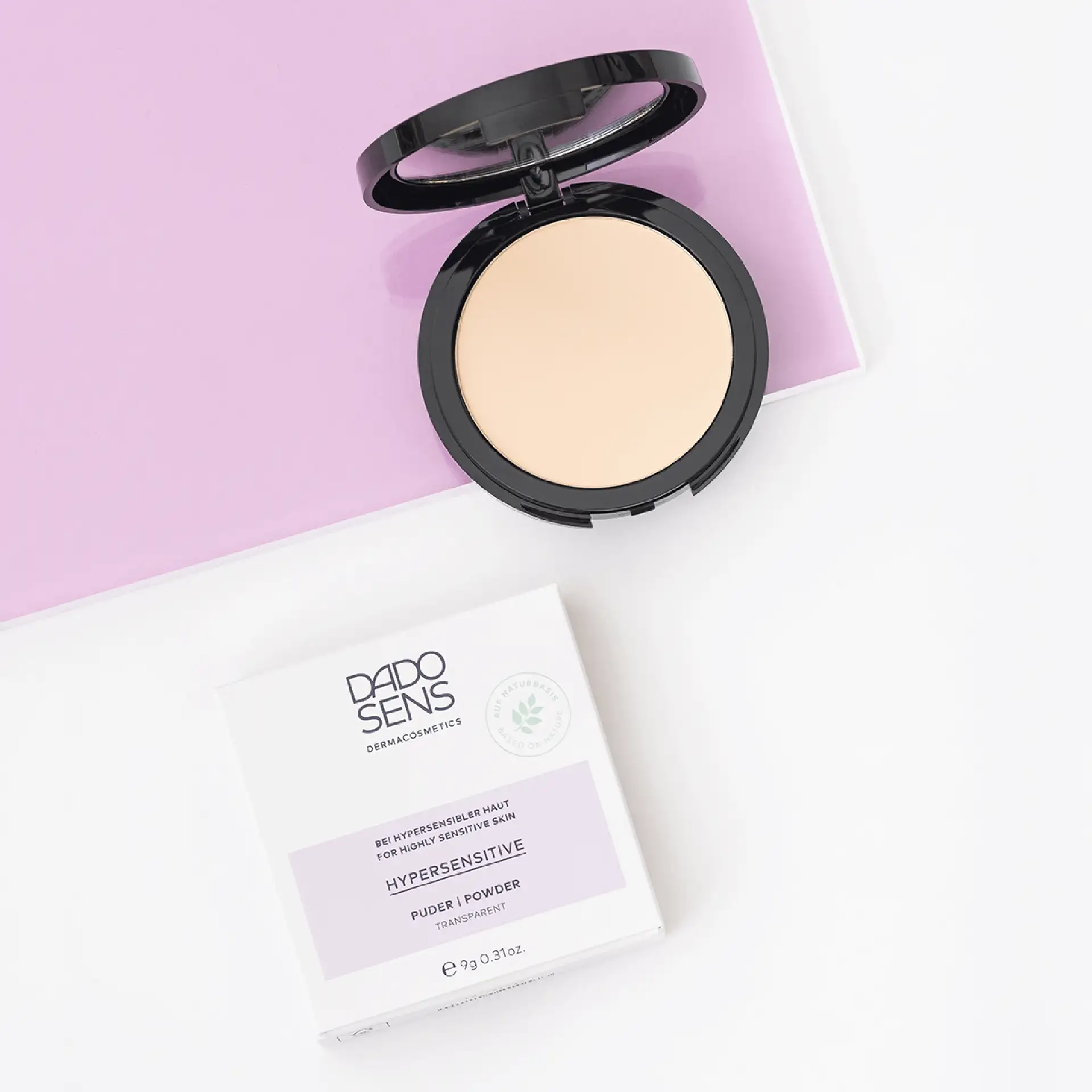 DADO SENS Hypersensitive transparenter Puder in schwarzer Dose mit Verpackung auf weiß-lila Untergrund