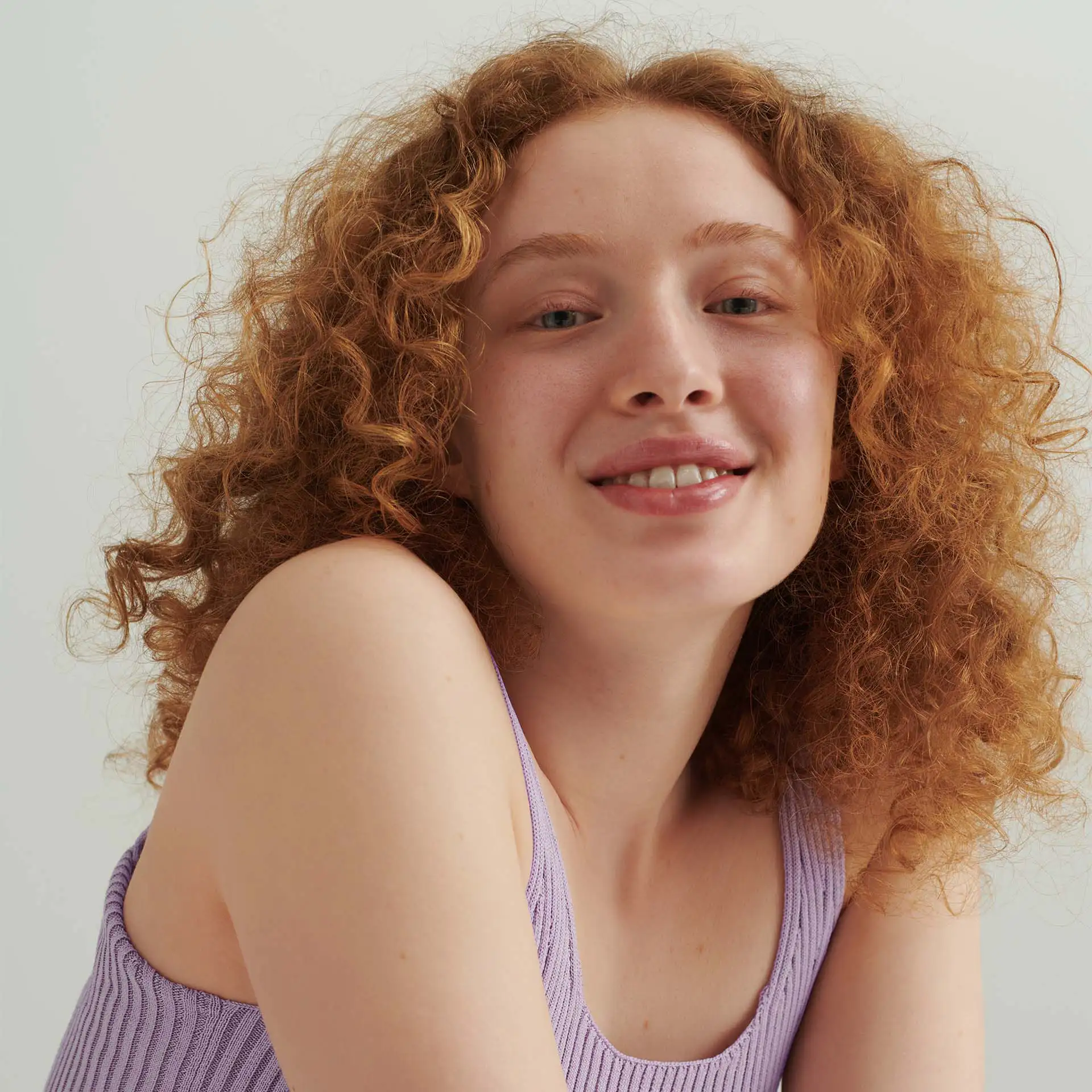 Nahaufnahme eines lächelnden Models mit roten Locken und lila Top vor hellem Hintergrund