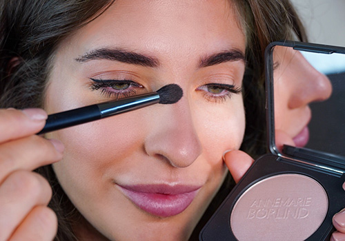 Frau trägt ANNEMARIE BÖRLIND Coral Glow Highlight-Puder mit Pinsel auf Nasenrücken auf, Spiegel sichtbar