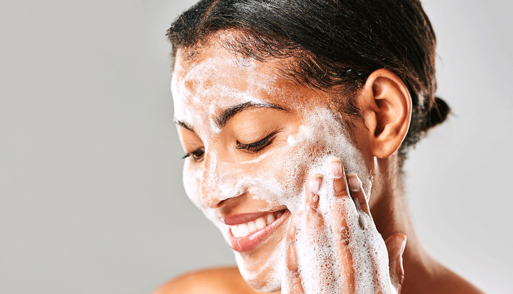 Frau wäscht Gesicht mit Schaum, Augen geschlossen, entspanntes Lächeln, helle Hintergrundfarbe