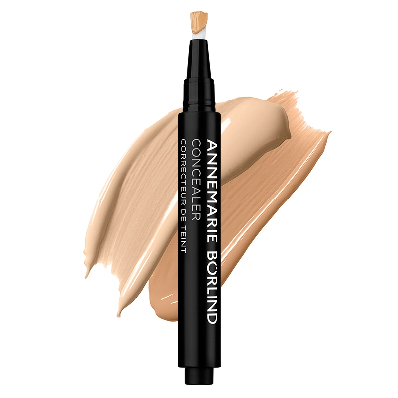 ANNEMARIE BÖRLIND Concealer in Stiftform mit Pinselapplikator, im Hintergrund zwei cremige Farbswatches in Beigetönen.