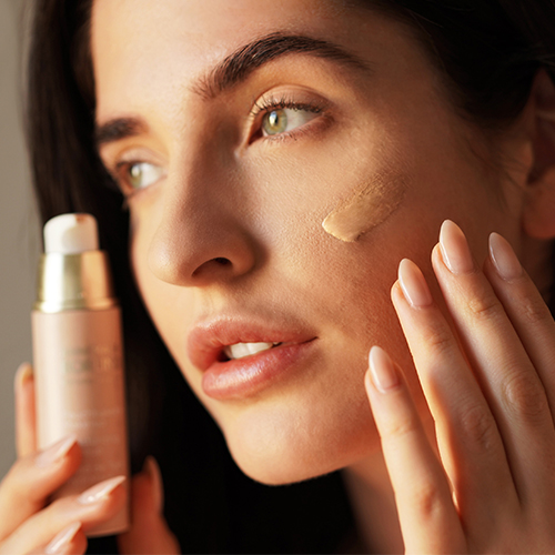 Frau trägt flüssige Foundation auf Wange auf, hält Pumpspender mit Foundation in der Hand