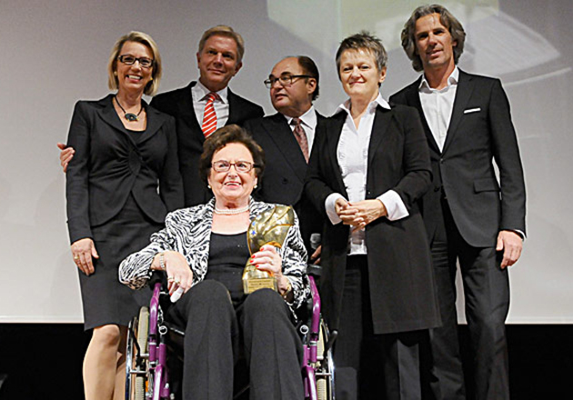 Fünf Personen in formeller Kleidung stehen hinter Annemarie Lindner im Rollstuhl, die eine Trophäe hält.