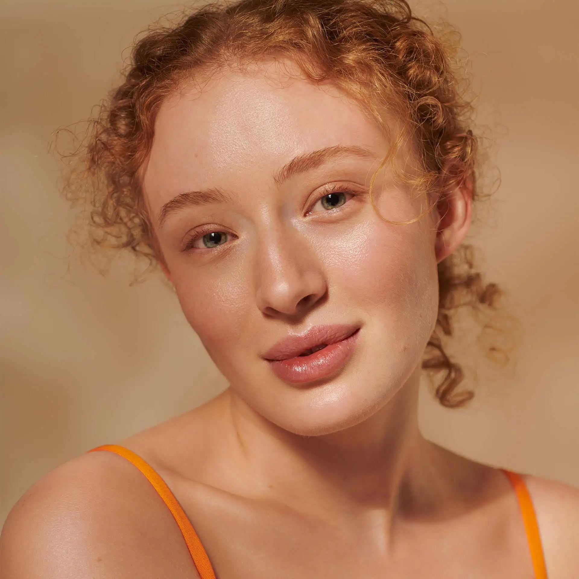 Nahaufnahme eines Models mit lockigem roten Haar und natürlichem, leicht glänzendem Hautbild vor beigem Hintergrund