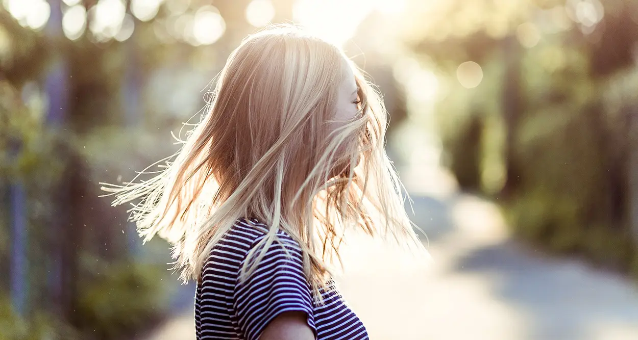 Frau mit langen blonden Haaren dreht sich im Sonnenlicht auf einem unscharfen Waldweg