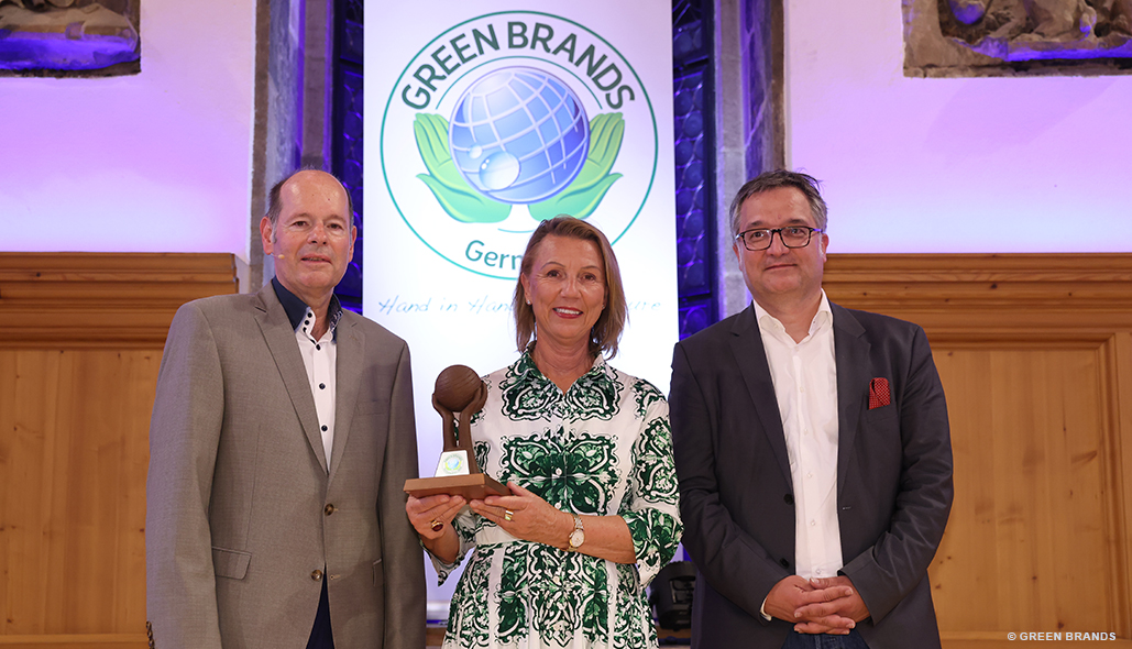 Drei Personen stehen vor GREEN BRANDS Germany Logo, Frau hält eine Trophäe in einem Veranstaltungsraum