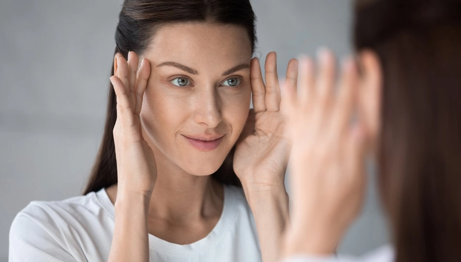 Woman looking in the mirror and gently massaging her eye area – anti-aging eye care tips