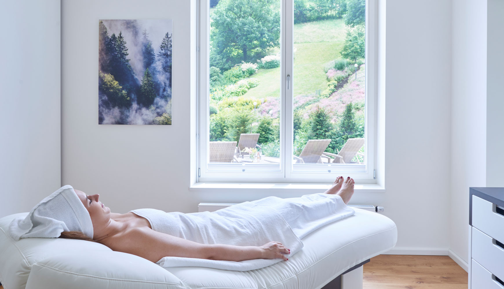 Frau entspannt auf Spa-Liege mit Handtuch, hellem Raum und Blick auf Garten mit Liegestühlen