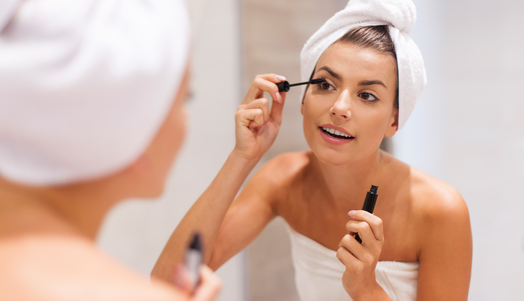 Frau mit Handtuch auf dem Kopf trägt Mascara auf ihre Wimpern vor einem Badezimmerspiegel auf