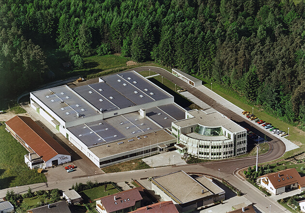 Großes Industriegebäude mit rundem Bürotrakt, umgeben von Wald und Wohnhäusern in ländlicher Gegend