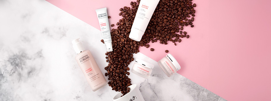ANNEMARIE BÖRLIND ENERGY NATURE Pflegeserie mit Creme, Lotion und Tiegeln, umgeben von Kaffeebohnen auf rosa Hintergrund