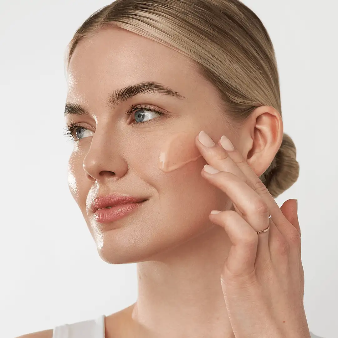 Frau trägt mit Finger eine rosafarbene Creme auf die Wange auf, Hautpflegeanwendung im Nahaufnahme