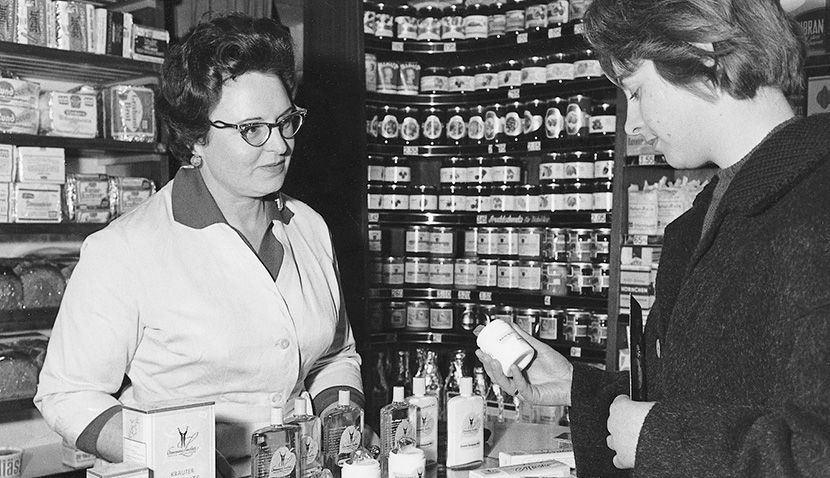 Apothekerin berät Kundin, die ein Produkt aus einer Auswahl von Cremes und Lotionen hält, Regal mit Gläsern im Hintergrund