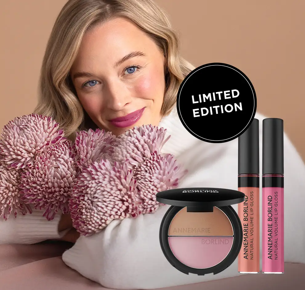 Frau mit ANNEMARIE BÖRLIND NATURAL HUG SET Make-up, lehnt sich lächelnd mit Blumenstrauß in Rosa- und Lilatönen auf Tisch vor beigem Hintergrund