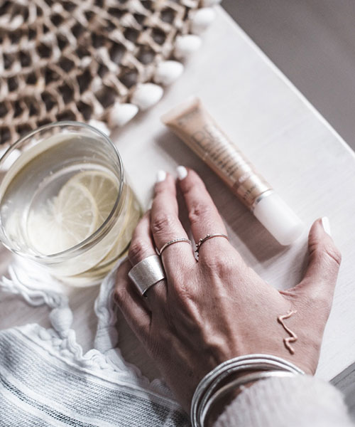 Hand mit silbernen Ringen und Armreifen neben Glas mit Zitronenscheiben und Hautpflege-Tube auf Tisch