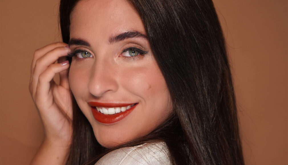 Nahaufnahme einer lächelnden Frau mit roten Lippen und grünlichen Augen vor braunem Hintergrund