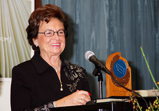 Annemarie Lindner steht lächelnd am Rednerpult mit Mikrofon und hält eine Auszeichnung aus Holz mit blauem Emblem