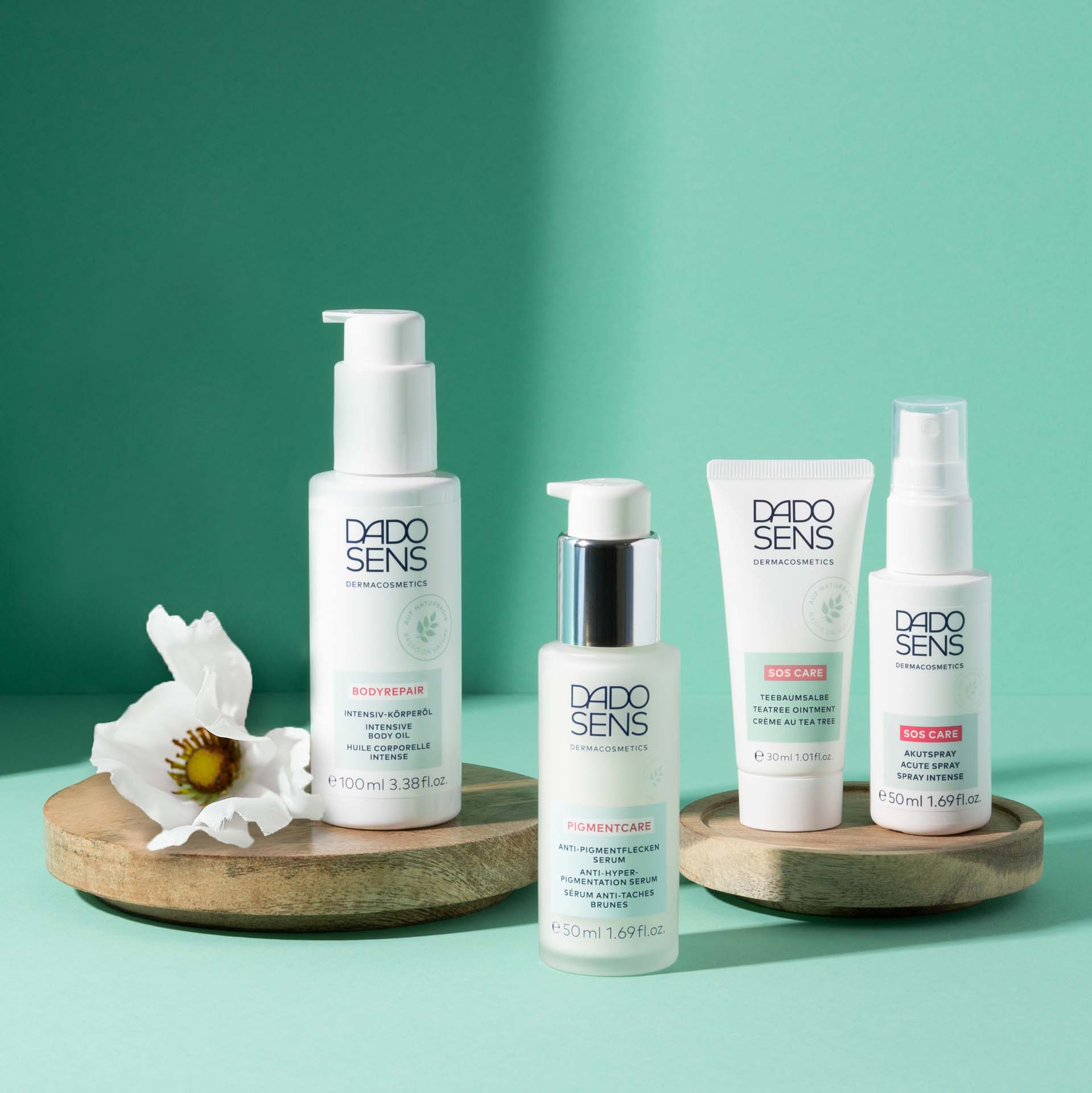 DADO SENS Intensiv-Körperöl, Anti-Pigmentflecken-Serum, Teebaumsalbe und Akutspray auf Holzplatten vor grünem Hintergrund