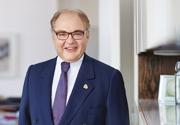 Mann im Anzug mit Krawatte und Brille steht lächelnd in einem hellen Büro mit Holzregal im Hintergrund