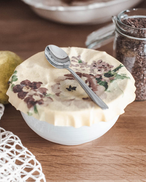 Schüssel mit floral bedachtem Stoffdeckel, Löffel darauf, daneben Glas mit Kakaonibs und Birne auf Holzfläche