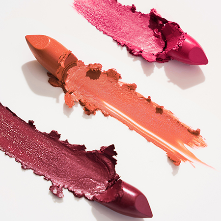 Drei Lippenstifte in Pink, Koralle und Dunkelrot mit Farbspuren auf weißem Hintergrund