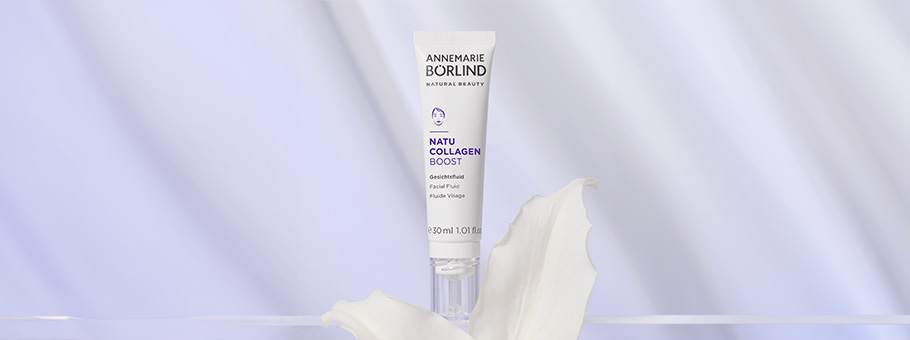 ANNEMARIE BÖRLIND NATU COLLAGEN BOOST Gesichtsfluid in weißer Tube vor hellviolettem Hintergrund mit weißer Blüte