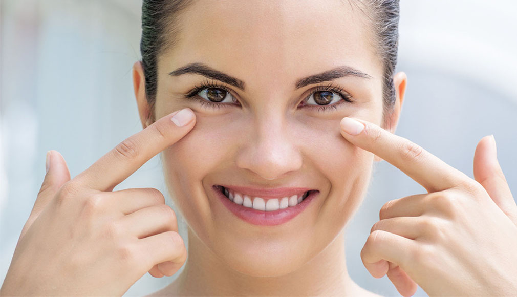 Anti-Aging-Augenpflege Frau zeigt mit den Fingern auf die Augenpartie und lächelt, helle, natürliche Beleuchtung