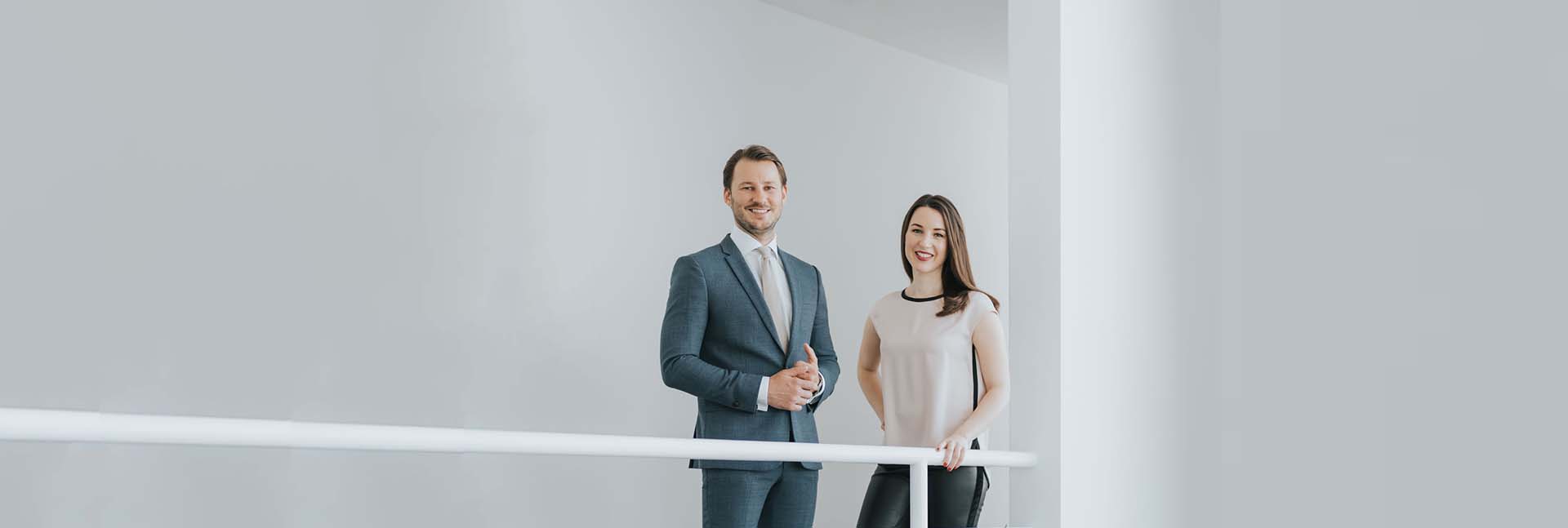 Zwei Personen in Businesskleidung stehen lächelnd an einem weißen Geländer in einem hellen, modernen Raum