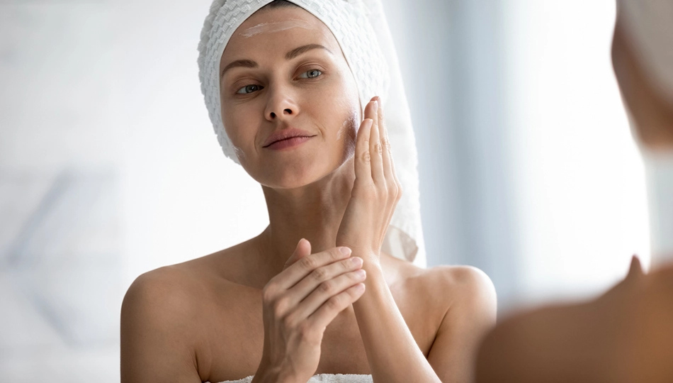 Gesichtspflege mit Hyaluronsäure für hydratisierte Haut Frau trägt Gesichtscreme mit Hyaluronsäure nach dem Duschen vor dem Spiegel auf