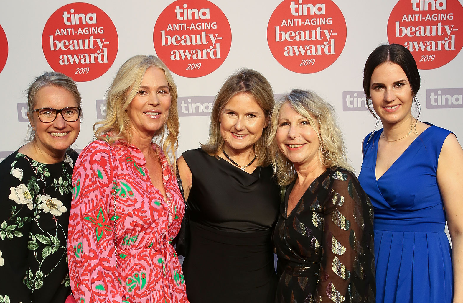 Fünf Frauen in festlicher Kleidung posieren vor einer Wand mit roten "tina ANTI-AGING beauty-award 2019" Logos