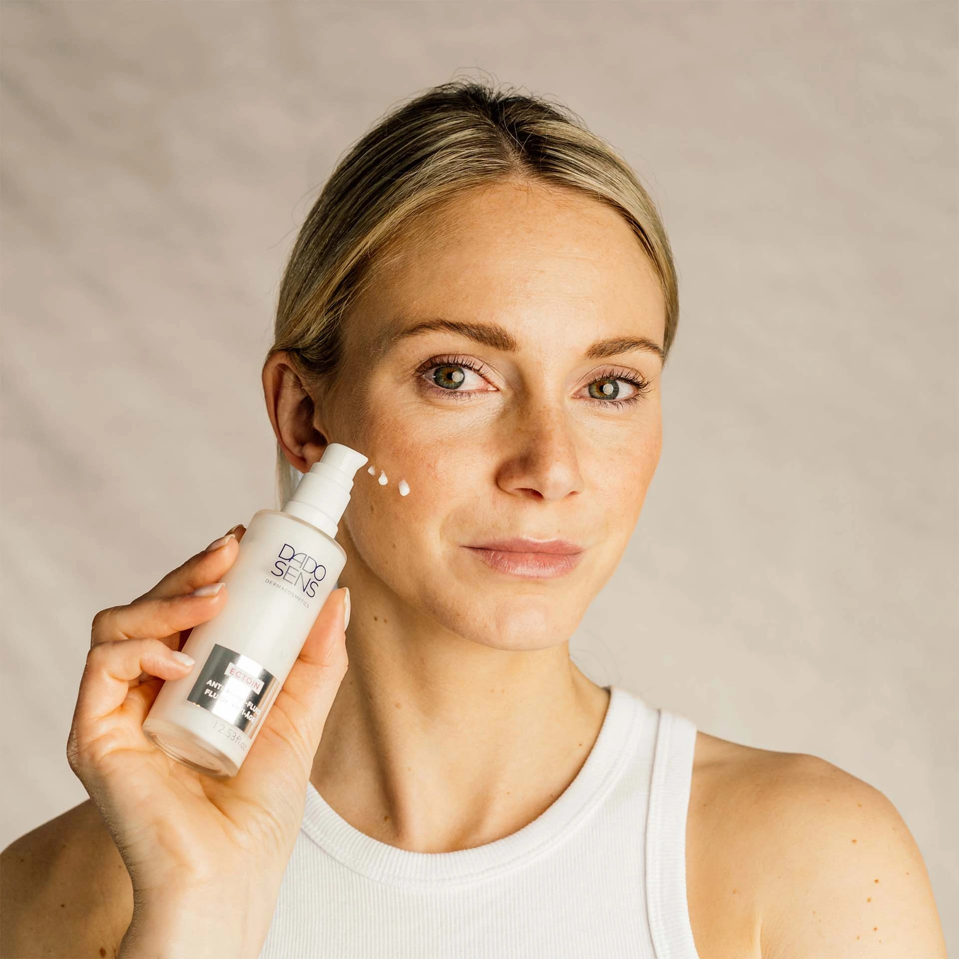 Frau trägt DADO SENS ECTOIN Spezialpflege auf Wange auf, hält Flasche mit Pumpspender vor hellem Hintergrund