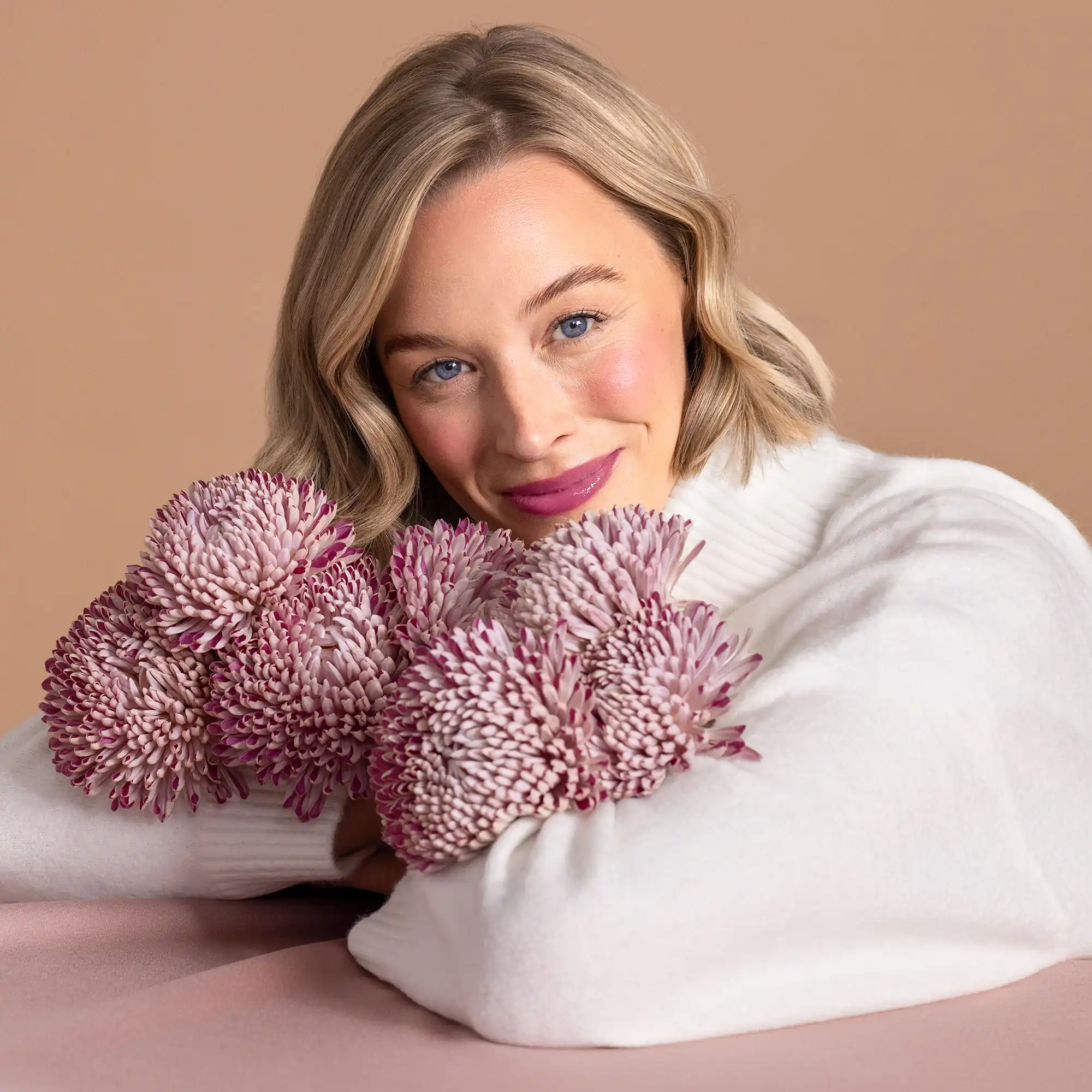 Frau mit ANNEMARIE BÖRLIND NATURAL HUG SET Make-up, trägt Bronzer, Blush und Lip Gloss in Farben Nude Hug und Snuggle Berry, lächelt mit Blumenstrauß in Rosa- und Lilatönen